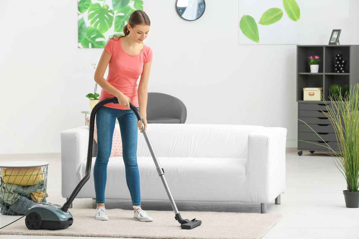 A woman is vacuuming the carpet in a living room. She is wearing a pink shirt and jeans. The room, featuring light-colored furniture and green plants, showcases the best carpet for pets and high traffic areas.