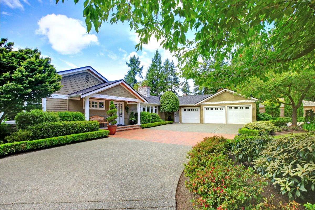 A well-maintained suburban house with a three-car garage, manicured lawn, and lush greenery, including shrubs and trees, under a clear sky. Perfect for families looking to know what to fix when selling a house.