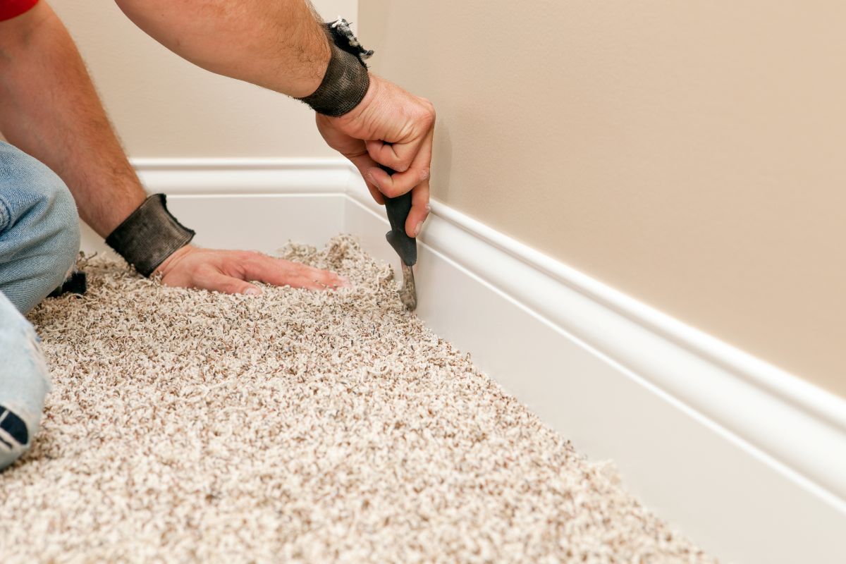 A person installing beige carpet along a white baseboard using a carpet tucking tool, an essential task when considering what to fix when selling a house.