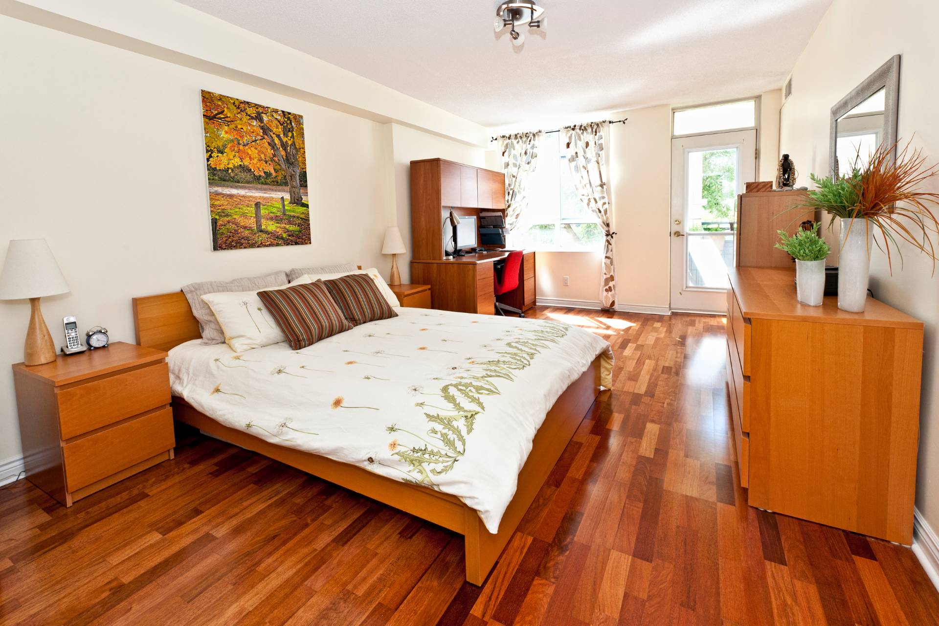 A new bedroom in a cozy home features a wooden bed frame, white and green bedding, two bedside tables with lamps, a desk, a dresser, a wall painting, a large window, and wooden floors.