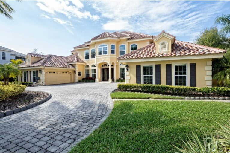 A large, yellow two-story house with a red tile roof and multiple arched windows, surrounded by a green lawn and driveway made of pavers showcases one of the best home exterior ideas.