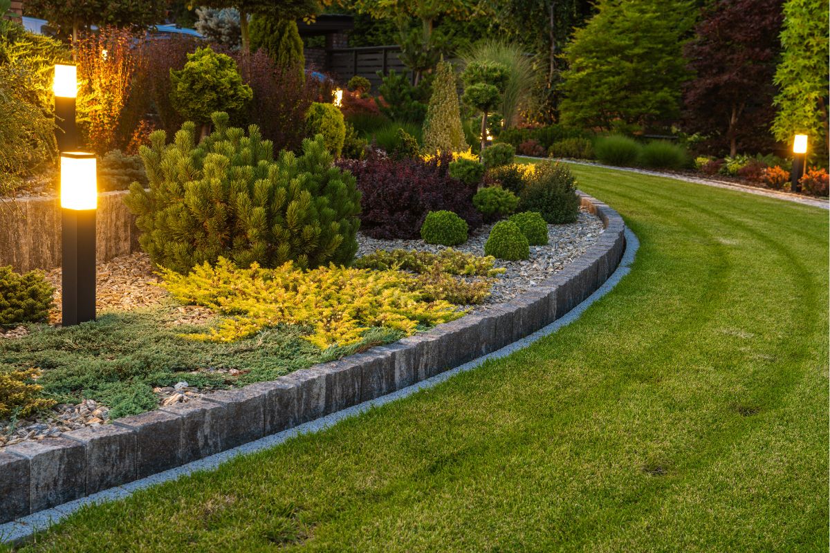 A well-maintained garden with an illuminated stone border, various shrubs, and a neatly mowed lawn in the evening offers excellent home exterior ideas.