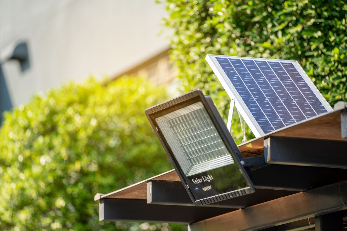 A solar-powered light fixture is mounted on a wooden structure, with a solar panel angled to capture sunlight, making it a perfect addition to your home exterior ideas. Green foliage and part of a building are visible in the background.