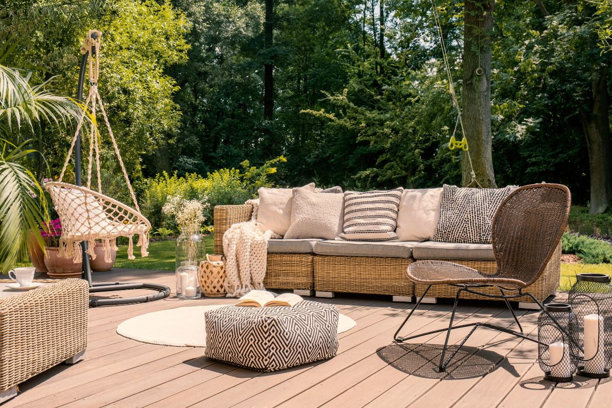 Outdoor patio with wicker furniture, including a couch with cushions, hanging chair, wireframe chair, and pouffe. Wooden decking adds to the cozy vibe, surrounded by greenery and trees — perfect for those seeking home exterior ideas.