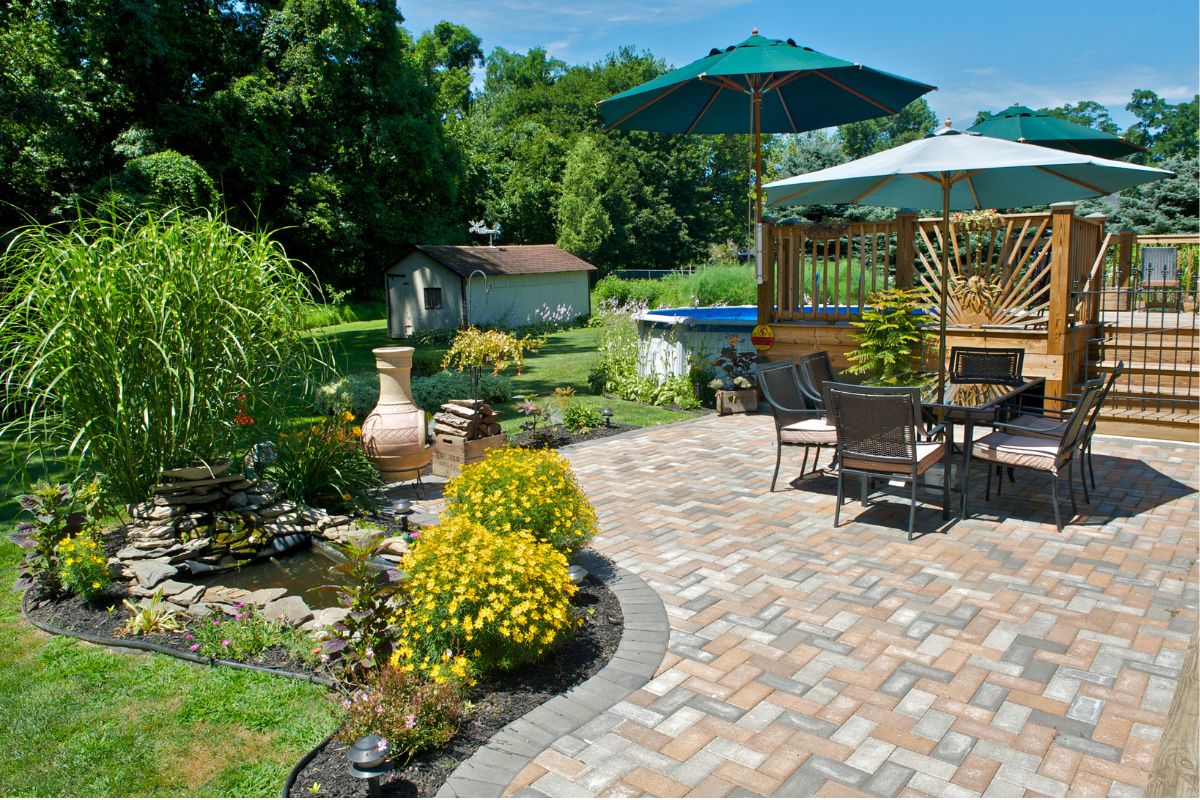 A backyard patio with a paved area featuring outdoor furniture and umbrellas. Adjacent is a landscaped area with plants, a small pond, and a shed visible in the background, offering perfect home exterior ideas.