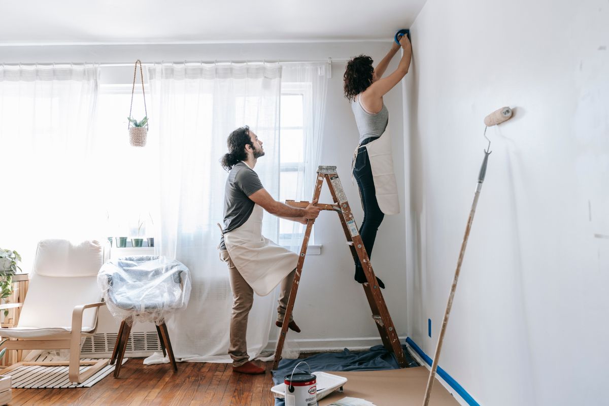 Two people are painting a room; one stands on a ladder painting the wall, while the other steadies the ladder. The room is covered in protective plastic sheets and painting supplies are on the floor—just part of what to fix when selling a house.