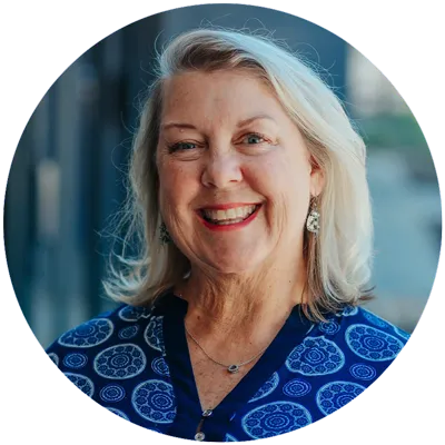 Portrait of a smiling woman with shoulder-length blonde hair, wearing a blue patterned top and earrings, standing in front of a blurred background. This description encapsulates her cheerful demeanor, making it perfect for an "About" section.