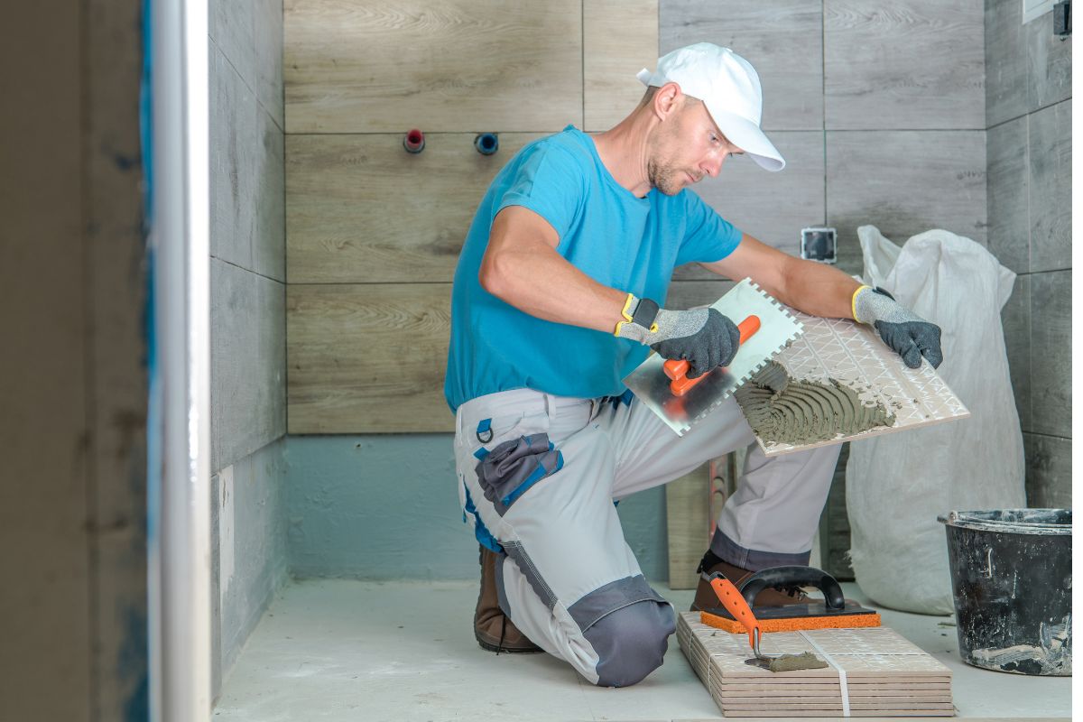 A worker in a blue shirt and white cap applies adhesive to a tile in a partially tiled room. He is kneeling on the floor, surrounded by tools and materials, addressing what to fix when selling a house.