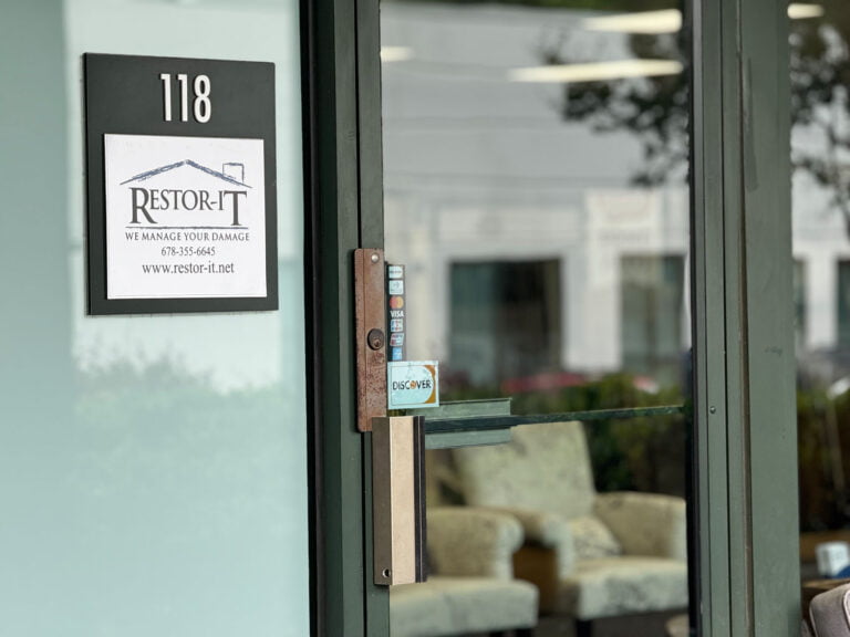 Glass door with a sign for "Restor-It" and entrance number 118. Inside, there are beige chairs and large windows reflecting outside greenery.