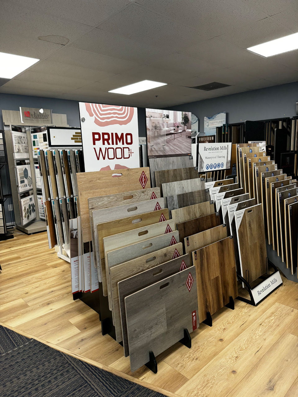 A store display featuring various wood flooring samples, including a prominent section labeled "Primo Wood+" with different shades and finishes available for customers to view.