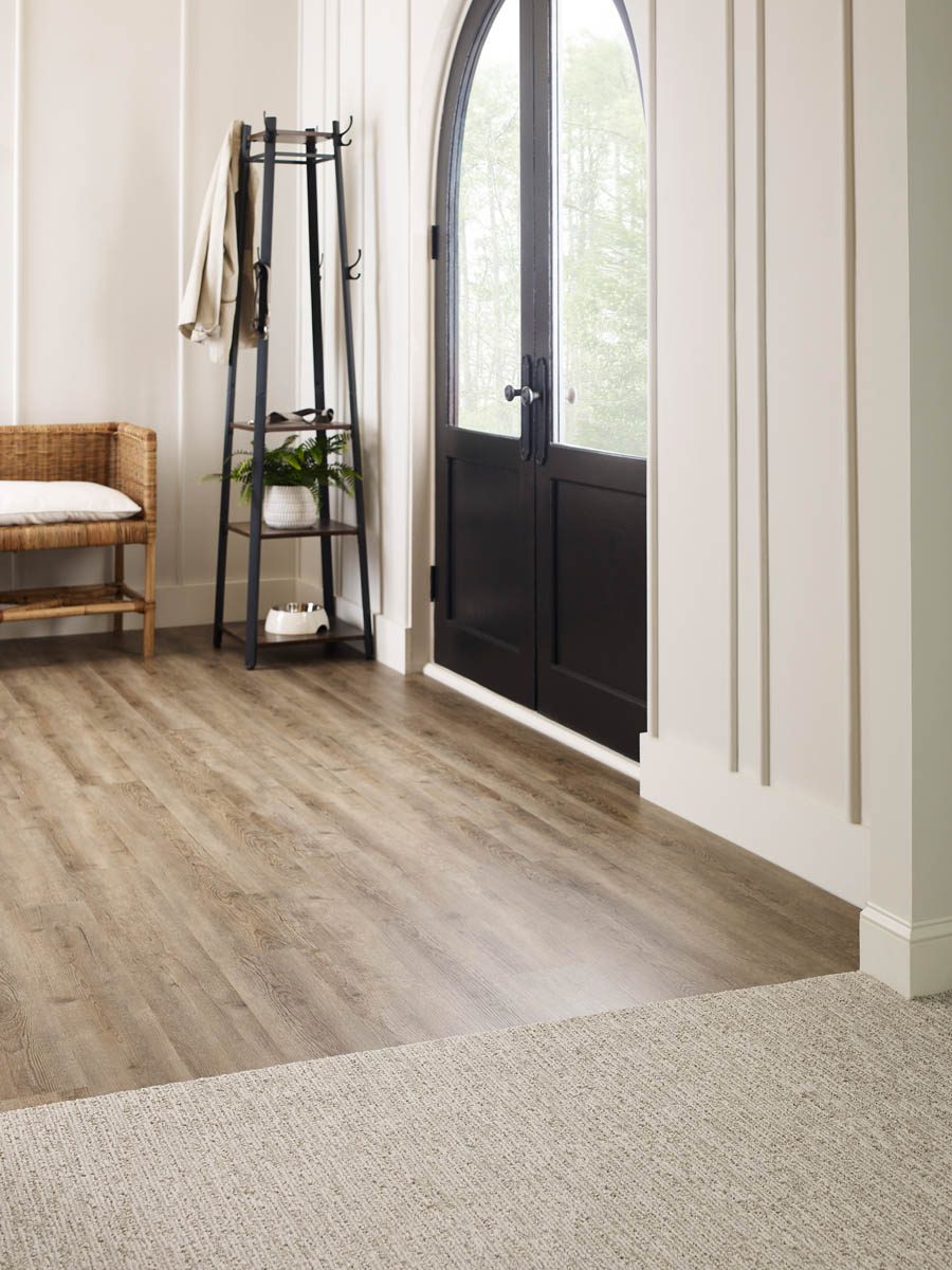 A minimalistic entryway featuring a wooden bench, black stand with coat and bag, wood and carpet flooring, and a black arched door with windows.