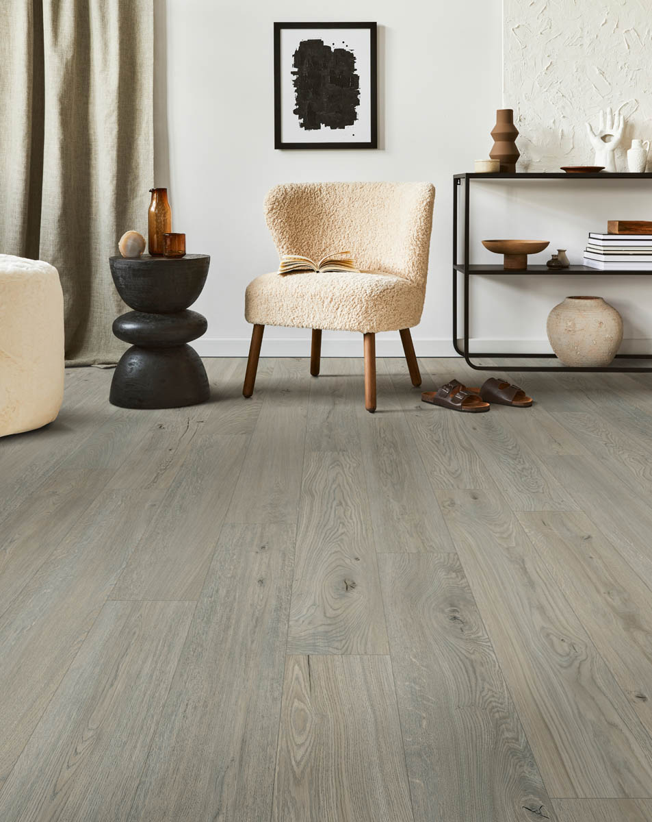 A modern living room features a beige armchair, a black side table, a wall-mounted abstract painting, and wooden flooring. Nearby, there is a shelf with decor items and a pair of brown slippers.