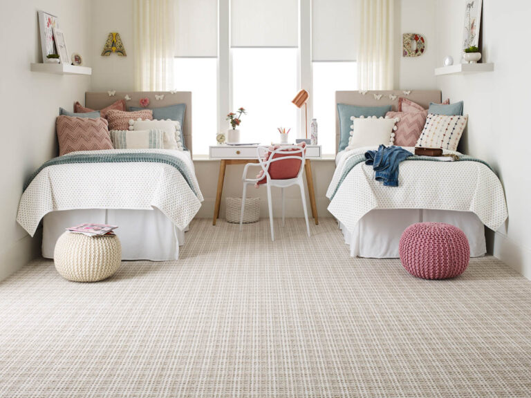 A symmetrical bedroom with two twin beds, a desk with a chair between them, decorative pillows, wall letters, and poufs on the floor. A large window in the center lets in natural light.