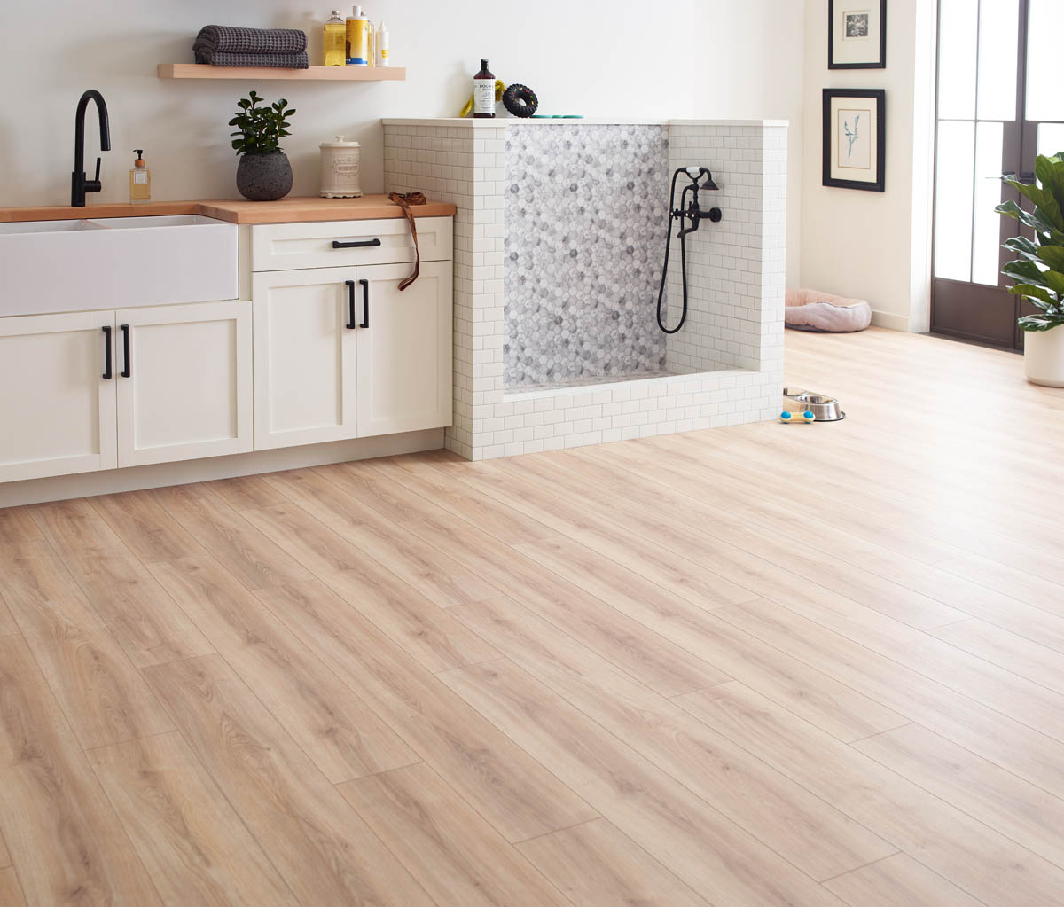 A laundry room with wooden flooring, a white farmhouse sink, white cabinets, wall shelves with plants and decor, and a pet washing station with a tiled backsplash and black faucet.