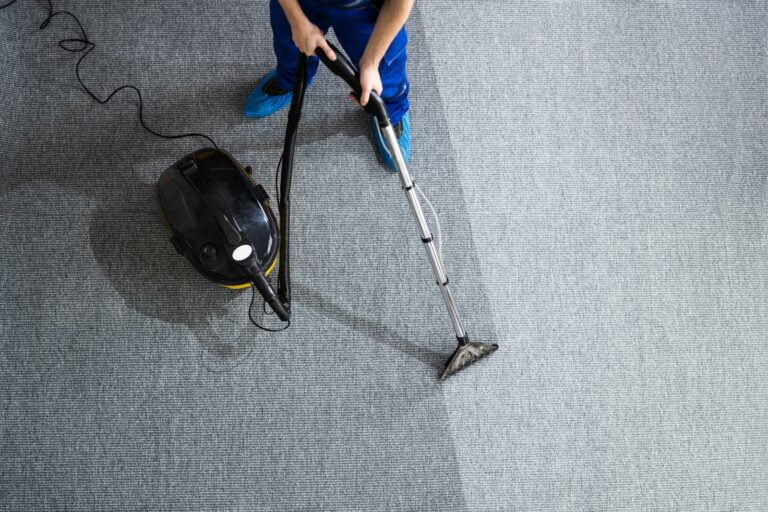 A person in a blue uniform and shoe covers operates a carpet cleaning machine, creating a visible clean path on the grey carpet. The professional ensures thorough carpet cleaning, restoring its original appearance.