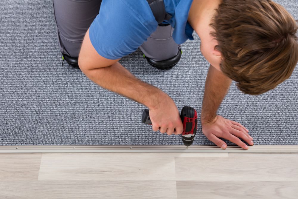 Person kneeling on a gray carpet, using a power drill to secure a strip at the edge where the carpet meets a wooden floor, showcasing skilled flooring installation services.