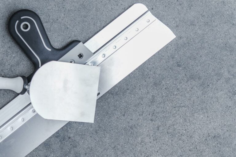 A metallic putty knife and a black-handled taping knife are placed on a gray concrete surface, ready for use in carpet stretching or patching tasks.