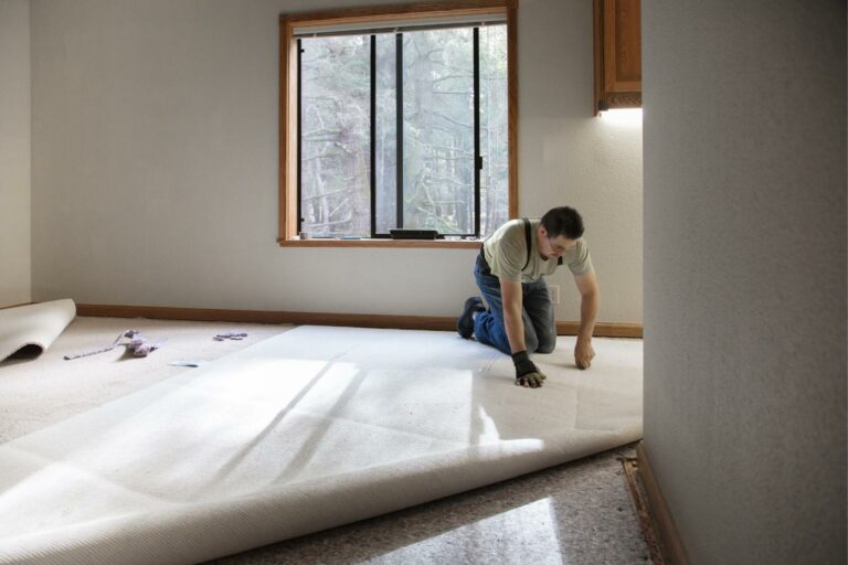 Person kneeling and installing carpet in a room with a large window. Tools are placed on the floor nearby, ready for any necessary carpet stretching adjustments.