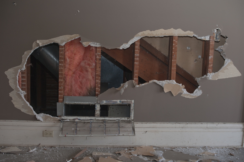 A room with a large hole in the wall shows exposed insulation, ductwork, and wooden beams. Debris is scattered on the floor, and a vent cover is partially detached at the bottom, indicating urgent need for repair services.