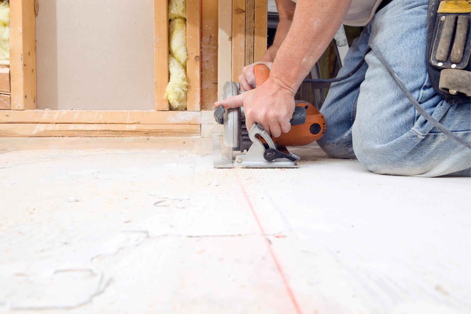 A person uses a circular saw to cut a wooden board on the floor, preparing for subfloor repairs. They are kneeling and wearing jeans with a tool belt on their waist.