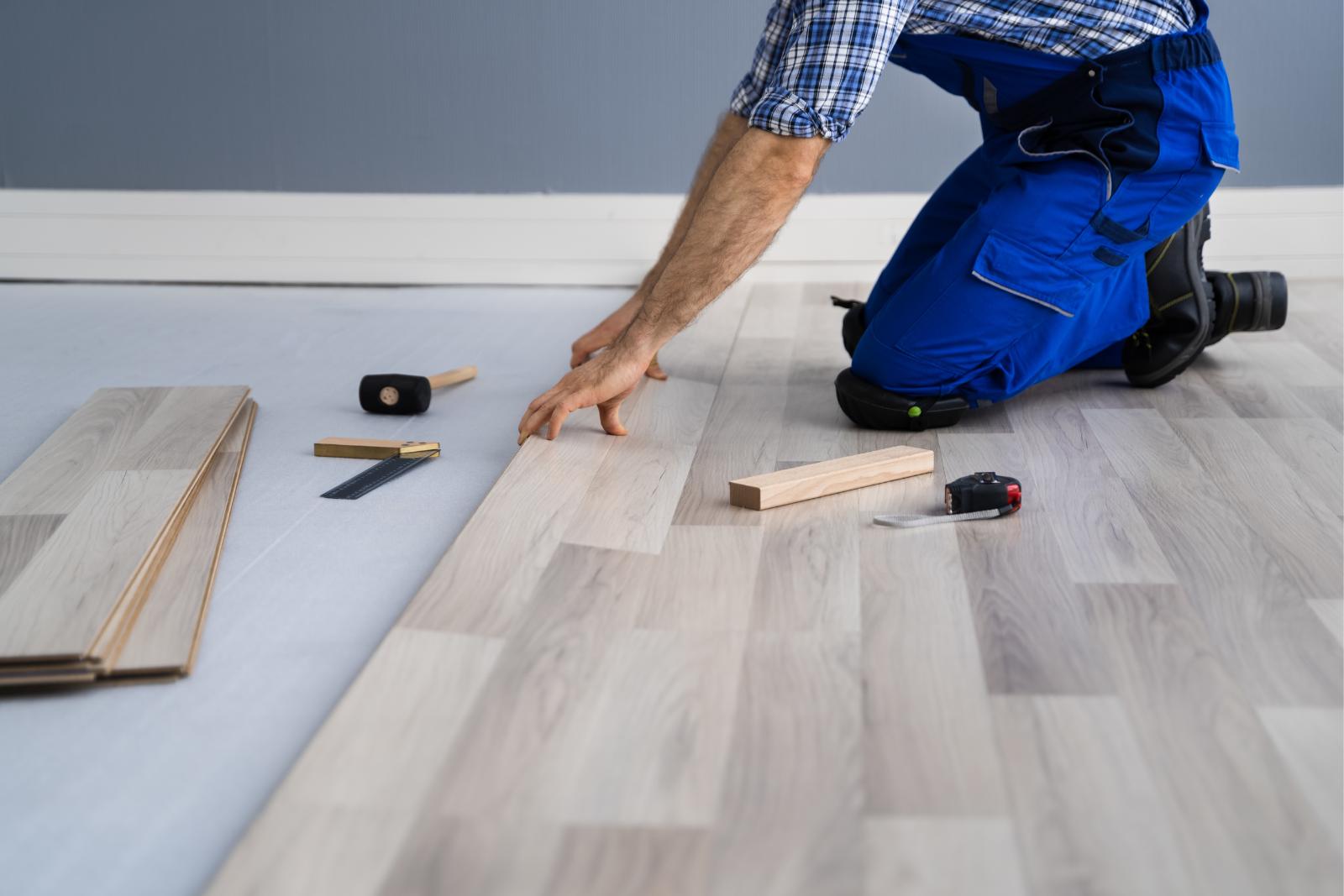 A person in blue overalls installs light wood flooring, surrounded by tools and plank pieces, carefully ensuring the subfloor is sturdy and level.