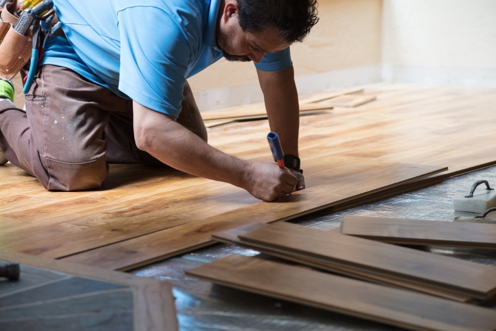 A person in a blue shirt installing wooden flooring, offering professional services as they measure and mark planks with a pencil while kneeling on the floor.