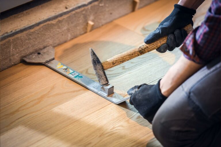 Person wearing gloves uses a hammer and a metal tool flooring and subflooring repairs, making sure the subfloor repairs are done meticulously.