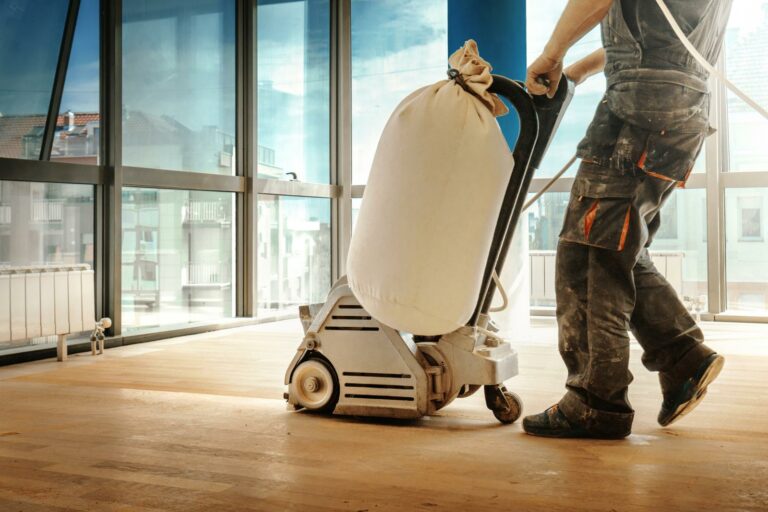 A person operates a large floor sanding machine in a sunlit room with large windows, ensuring the flooring is perfectly smooth before making any necessary repairs.