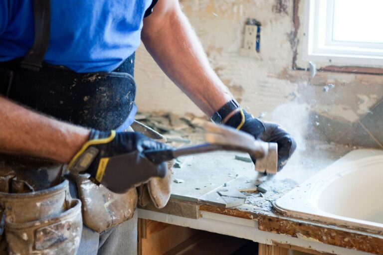 A person wearing a tool belt and gloves uses a hammer to remove tiles from a countertop next to a sink, diligently working on their home renovations.