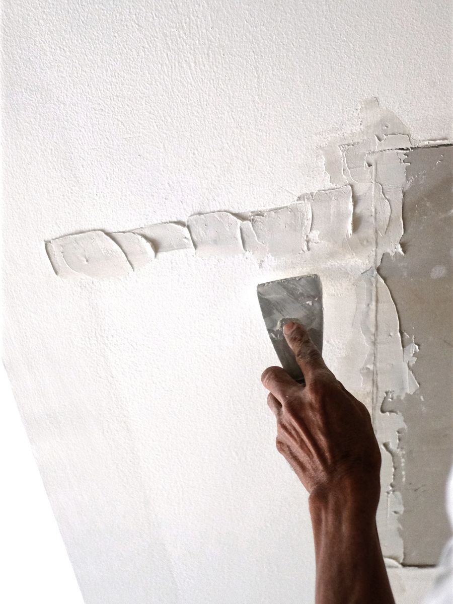 A person is using a putty knife to apply plaster on a white wall, expertly smoothing and repairing the surface in preparation for painting or drywall repair.