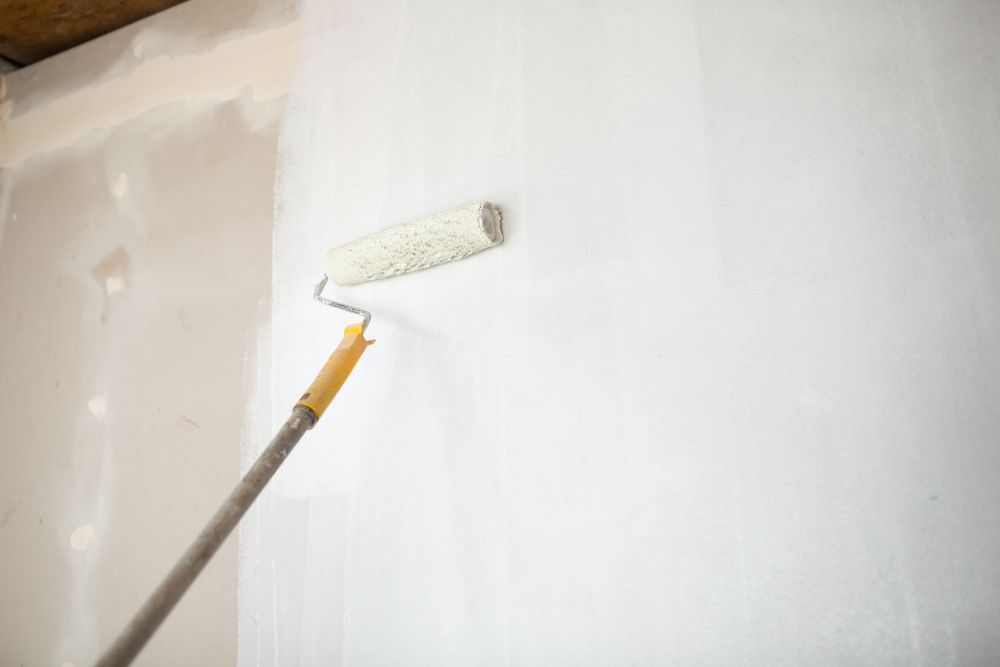 A paint roller with an extended handle is being used to efficiently tackle painting and drywall repair on a white wall.