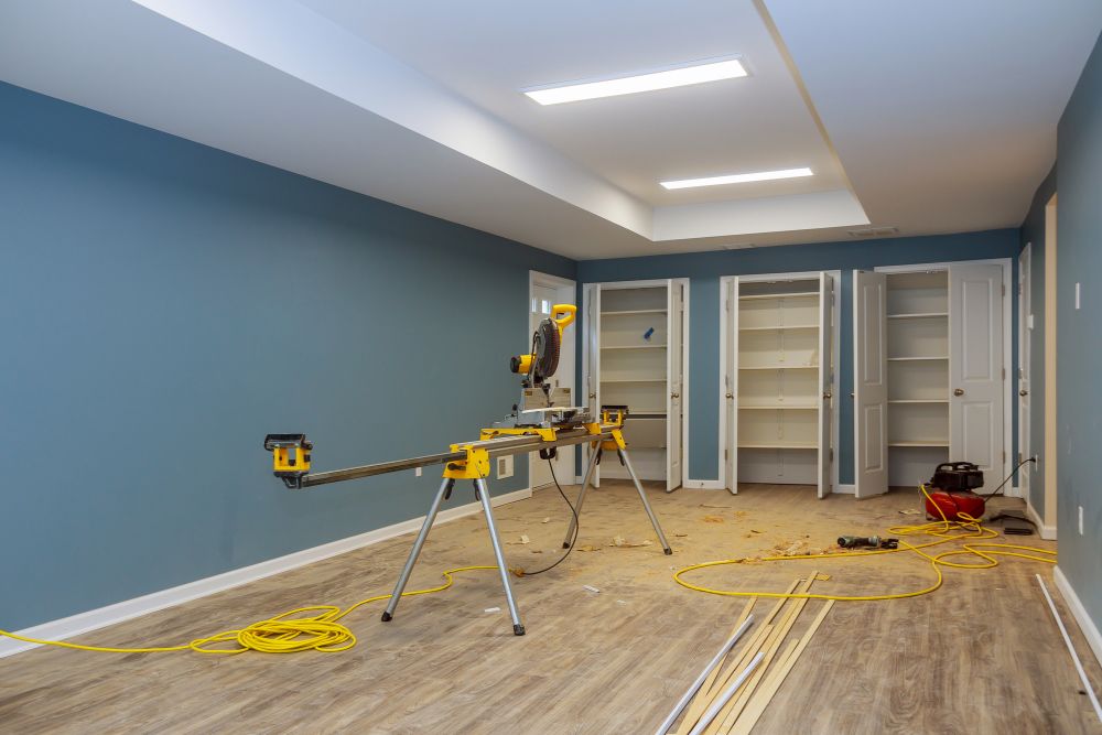 A room under renovation with blue walls, new wooden flooring, and open white closets. A miter saw on a stand, tools, and wooden planks are scattered on the floor, hinting at ongoing painting and drywall repair.