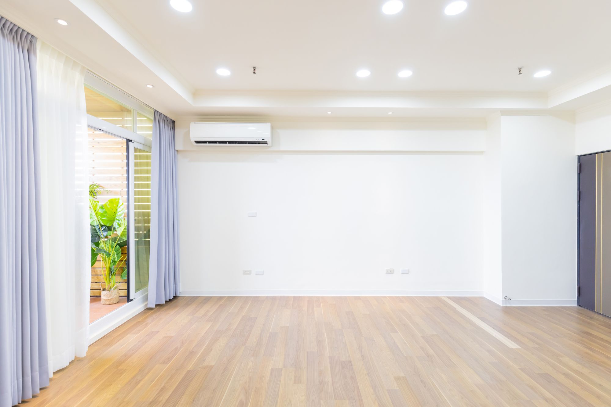 An empty room with wooden flooring, white walls, recessed lighting, and an air conditioning unit. Large windows with sheer curtains reveal a small outdoor area with potted plants. The space is perfect for painting and drywall repair projects.