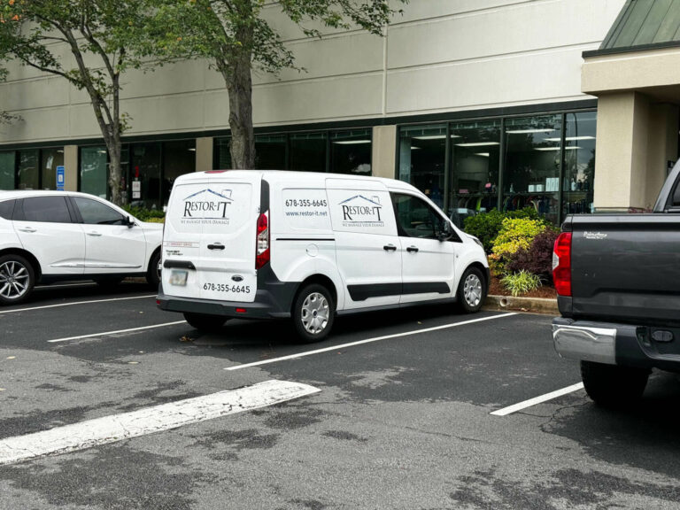 A white van with the "Restore-It" logo is parked in a space in front of a building. The van's contact information and website are about visible on its side and back. Other vehicles are also parked nearby.