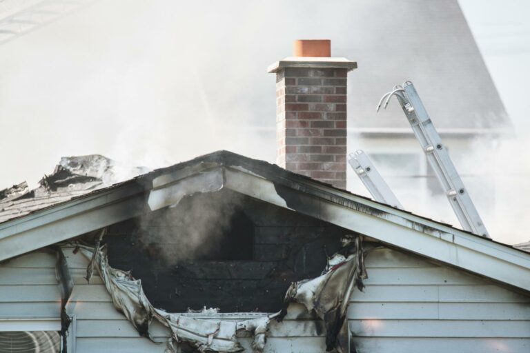 Close-up of a house with significant fire damage to the roof and siding. Smoke is rising from the burned area, and an emergency ladder is propped against the house's exterior, awaiting restoration efforts.