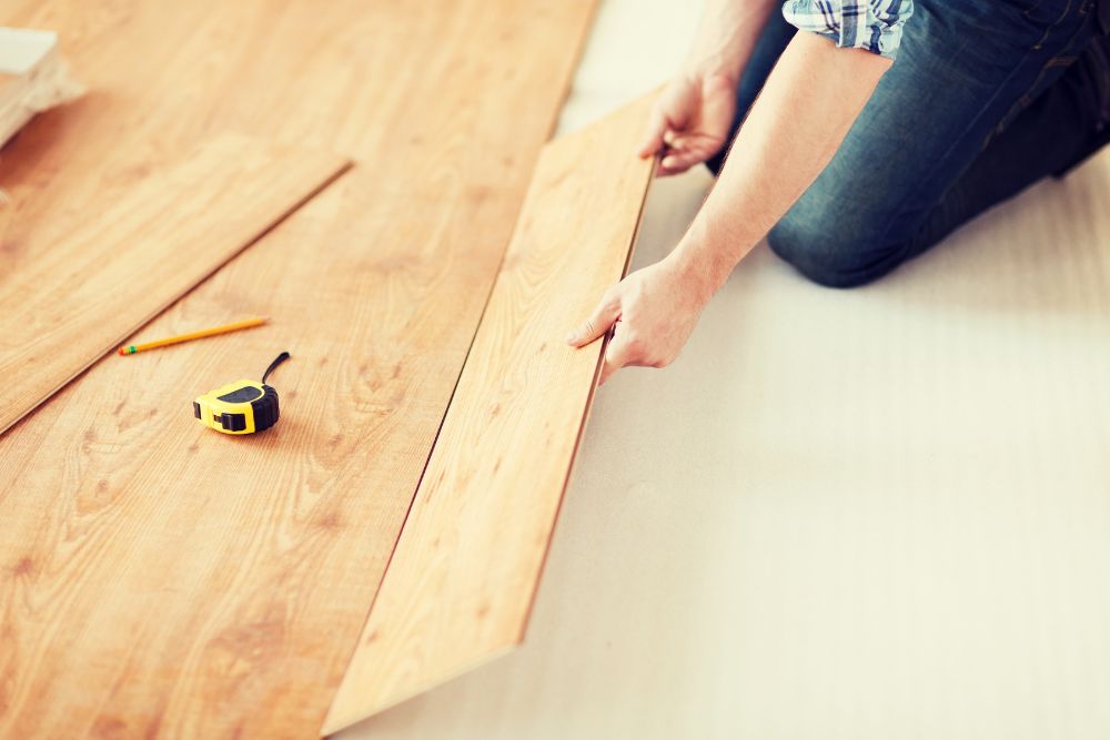 A worker from the Renovation and Restoration Company, dressed in jeans and a plaid shirt, is installing laminate flooring while holding a plank of wood. A pencil and tape measure are placed on the partially installed floor.