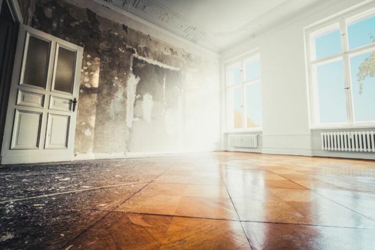 A partially renovated room displays a weathered wall and a bright, clean section with large windows, white walls, and polished wood flooring. The craftsmanship of our Renovation and Restoration Company is evident in the seamless blend of old and new, further accentuated by an open white door.