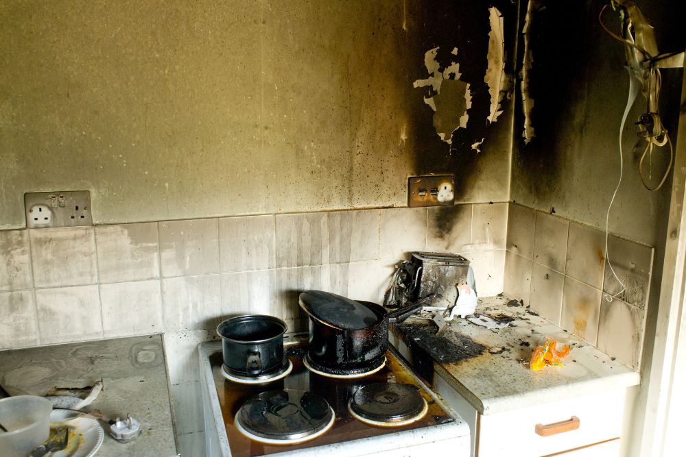 A kitchen with fire damage shows blackened walls, burnt appliances, charred pots on the stove, and partially melted kitchen surfaces, awaiting transformation by a skilled Renovation and Restoration Company.