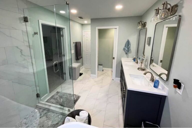 A modern bathroom, expertly crafted by our Renovation and Restoration Company, features a glass-enclosed shower, double sink vanity, marble floor tiles, and a doorway leading to another room.