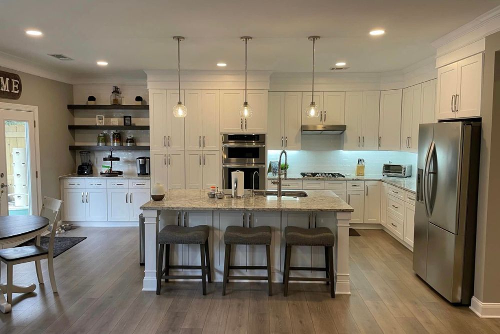 Modern kitchen with white cabinets, stainless steel appliances, and a kitchen island with three gray barstools. The island features a sink, and pendant lights hang above. A round table and chairs are on the left. Expertly crafted by our renowned Renovation and Restoration Company.