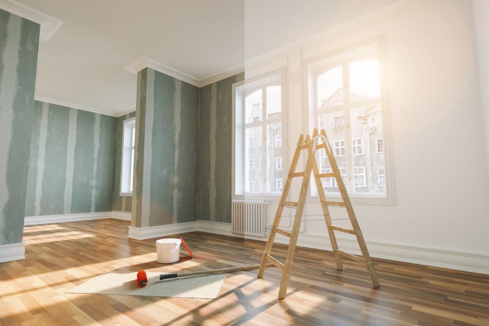 A sunlit room with hardwood flooring, partially painted striped walls, a ladder, a paint roller, and a paint bucket on a protective sheet in the corner; it's clear that the Renovation and Restoration Company is hard at work.