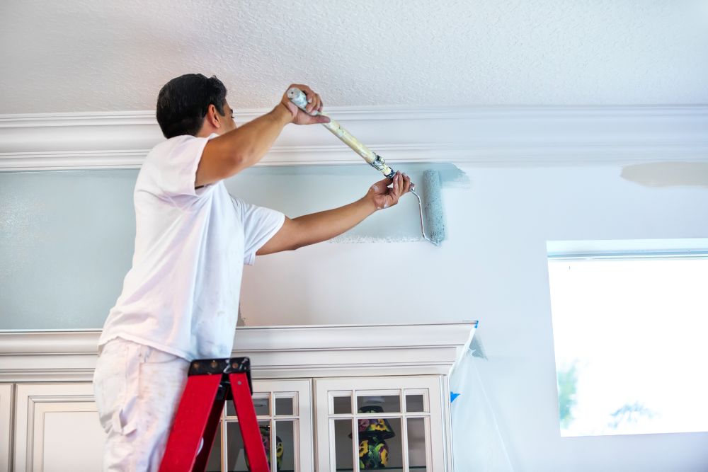 A person stands on a red ladder, painting a wall light blue with a roller brush. The wall, featuring white trim and a partially visible window, is undergoing not just painting but also some drywall repair to ensure a flawless finish.