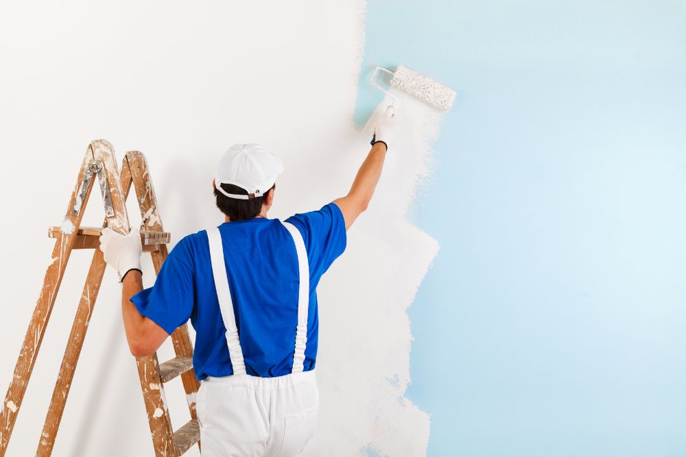 The man in a blue shirt and white overalls is focused on painting the wall with a roller, transitioning from blue to white. A wooden ladder stands next to him, ready for his next task of drywall repair.