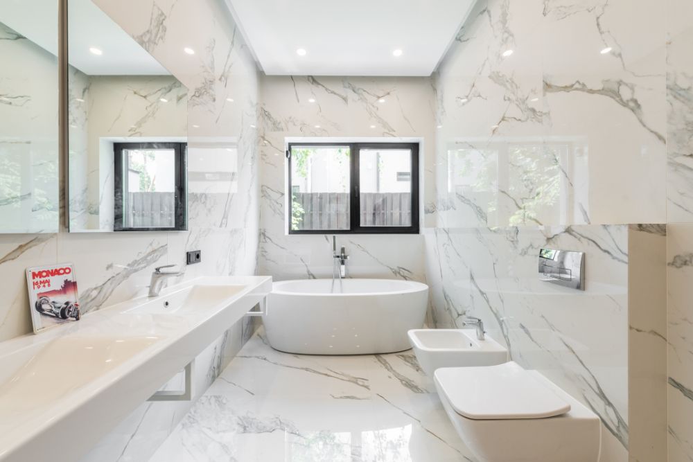 A modern bathroom with marble walls and floor, featuring a bathtub, double sink, bidet, and toilet. The room has a large mirror, windows, recessed lighting, and boasts resurfacing services to maintain its pristine condition.