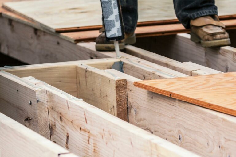 A construction worker applies sealant to the edge of a wooden subfloor, ensuring the structure is ready for flooring and any necessary repairs.