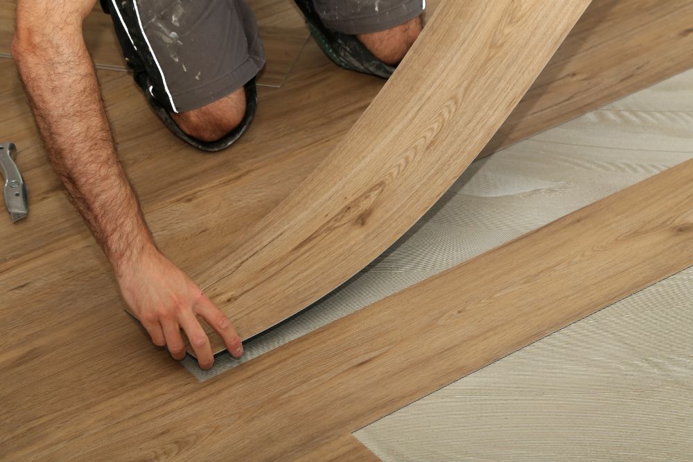 A person is installing a wooden floor panel by fitting it into place over a gray underlayment, showcasing their professional flooring installation skills.