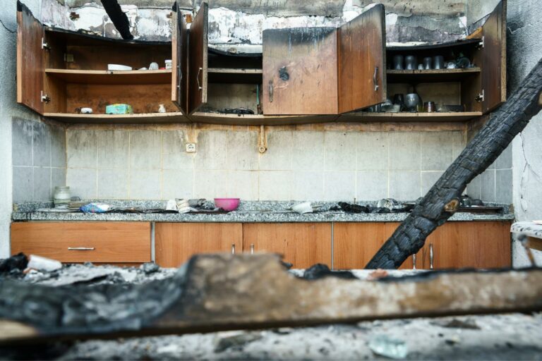 A damaged kitchen with burnt cabinets, scattered debris, and charred wood following a fire. Some shelves have remnants of kitchen items. The wall and countertops are stained from smoke and fire damage, compounded by water damage restoration efforts to salvage what remains.