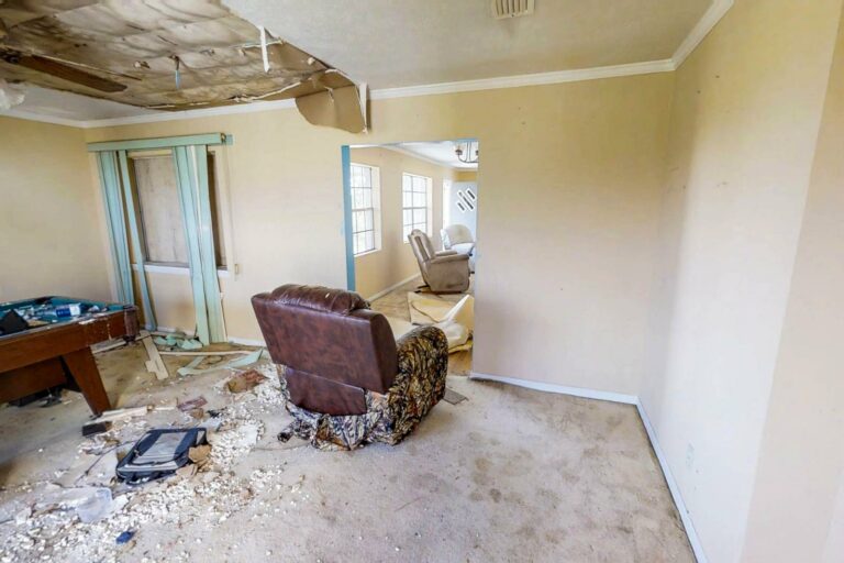 A room with a damaged ceiling, debris on the floor, and worn-out furniture. A pool table and books are in the corner, and the walls and carpet show signs of disrepair, likely in need of water mitigation after past damage.