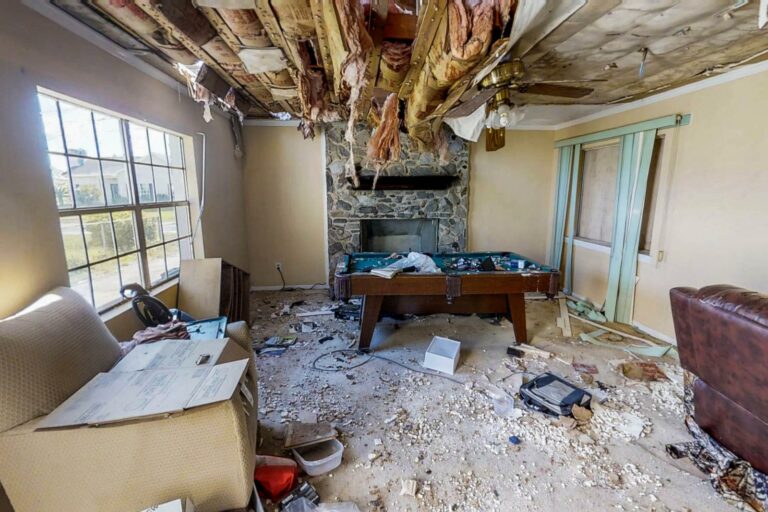 A living room with a severely damaged ceiling, debris scattered on the floor, and a pool table in the center. Signs of neglect mark the walls and furniture. A window lets in natural light, hinting at the urgent need for water mitigation to restore the space's former charm.