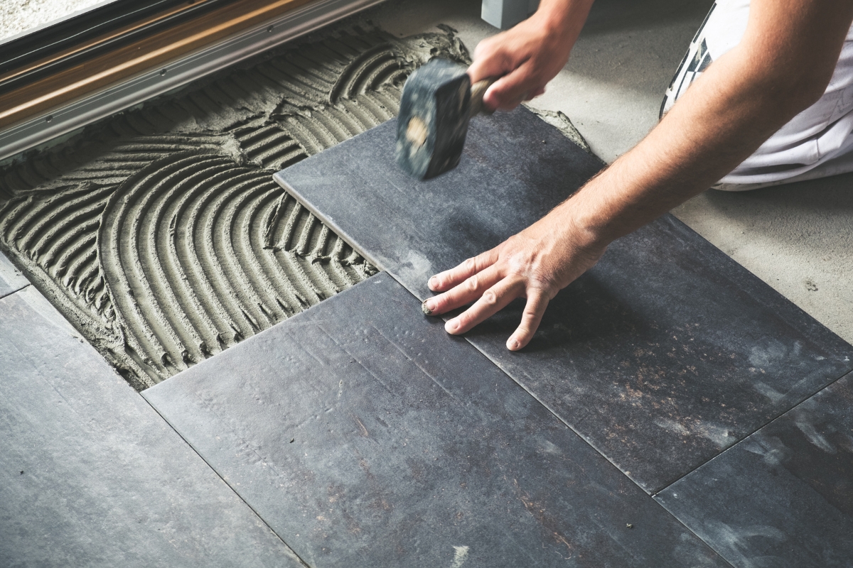 Person laying dark tiles on adhesive mortar in the bathroom subfloor, using a rubber mallet and their hand to position the tile correctly.